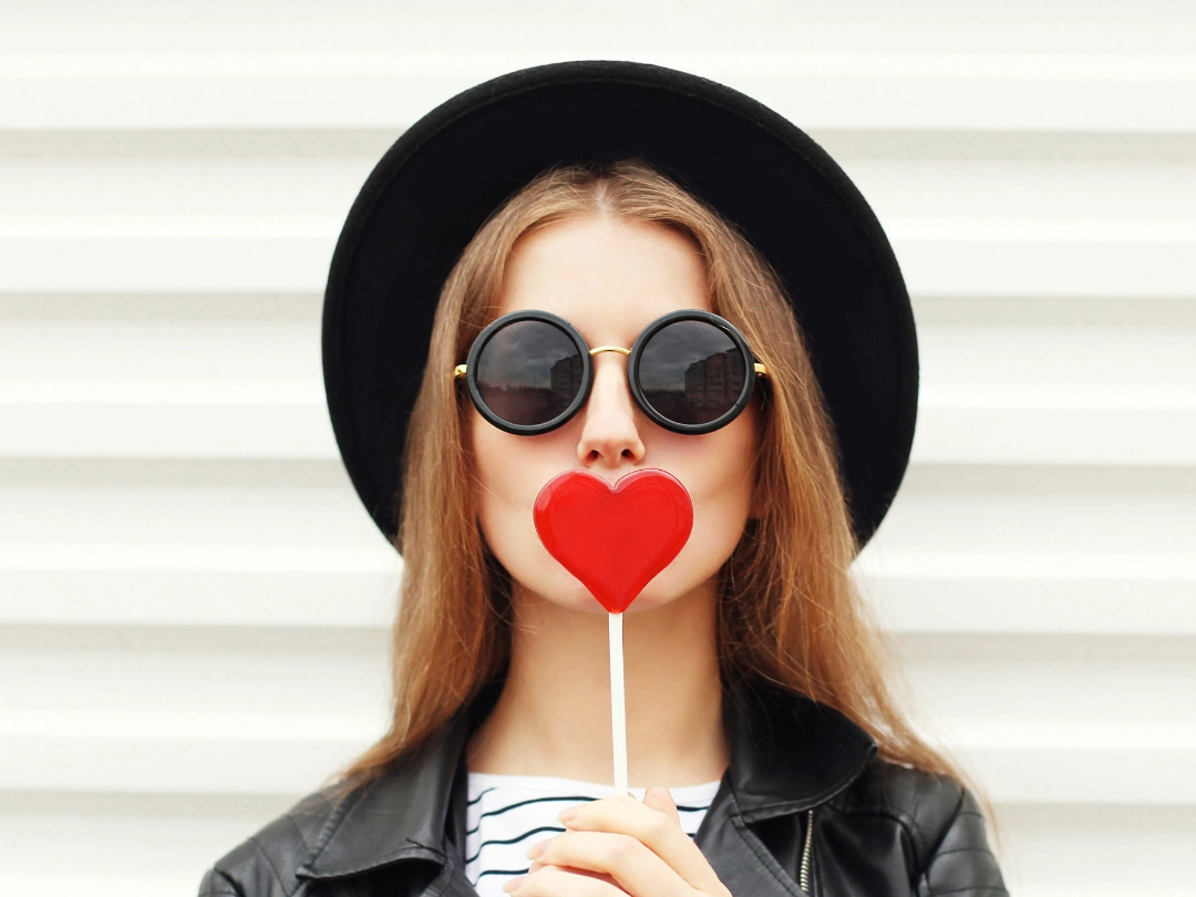 Junge Frau mit Sonnenbrille hält sich einen Lolly in Herzform vor den Mund.