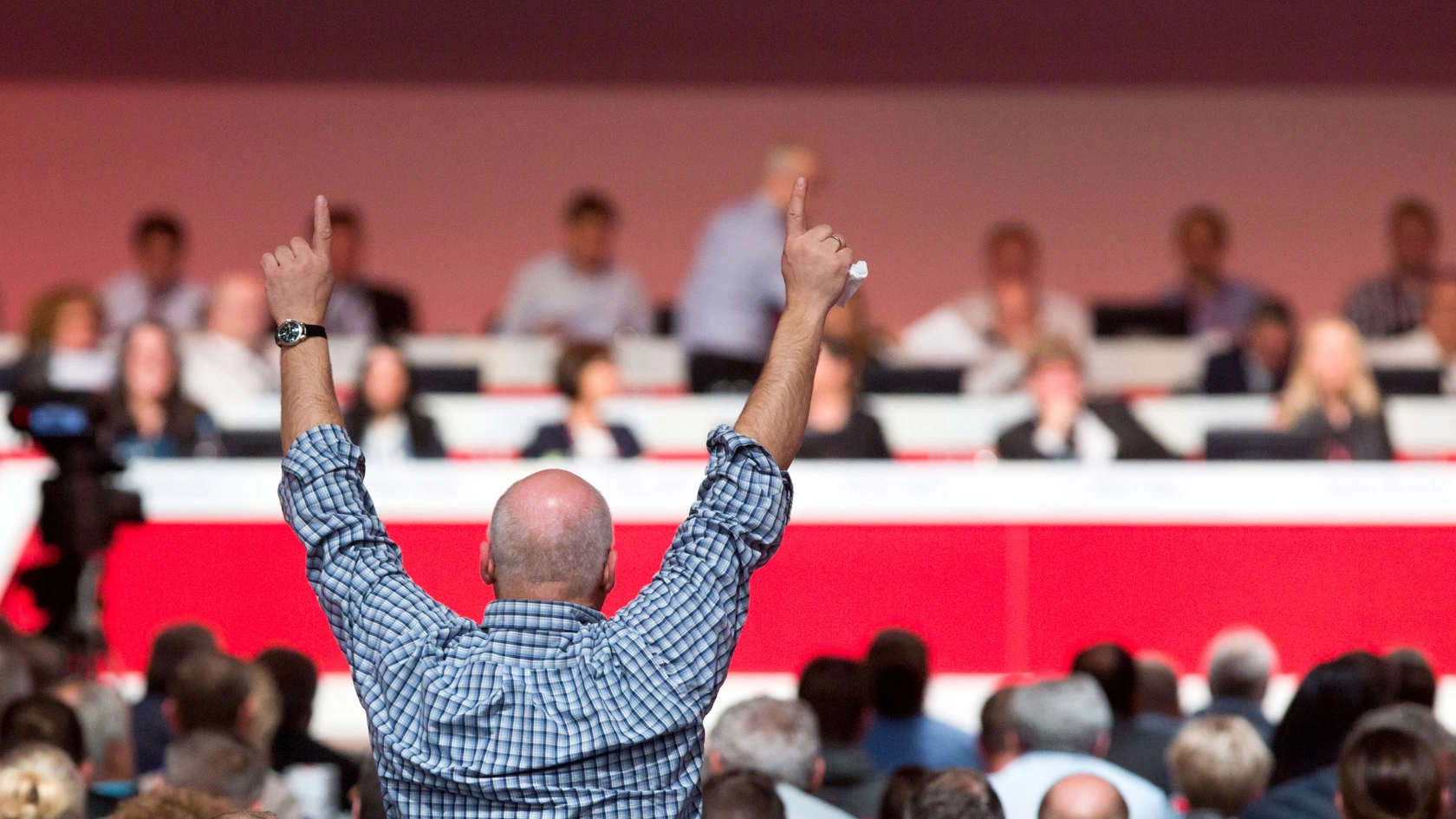 Mann im karierten Hemd steht während einer Konfernez mit beiden Händen nach oben auf.
